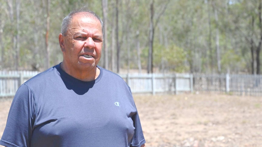 An aboriginal man wearing a blue shirt stands, serious with a dry brown grass covered field in the background.