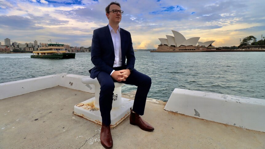 Man sits on a pier with a ferry and the Sydney Opera House in the background.
