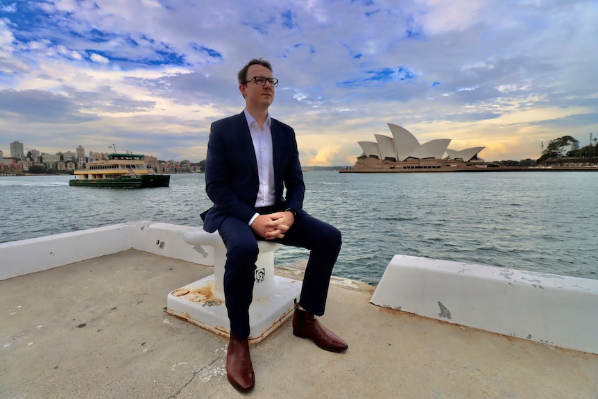 Man sits on a pier with a ferry and the Sydney Opera House in the background.