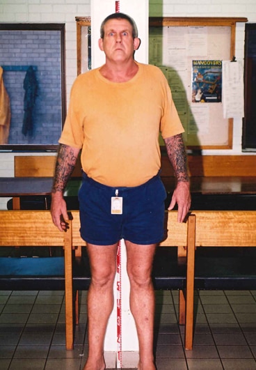 A large man in a yellow shirt and short blue shorts with tattoos on his forearms and bulging eyes
