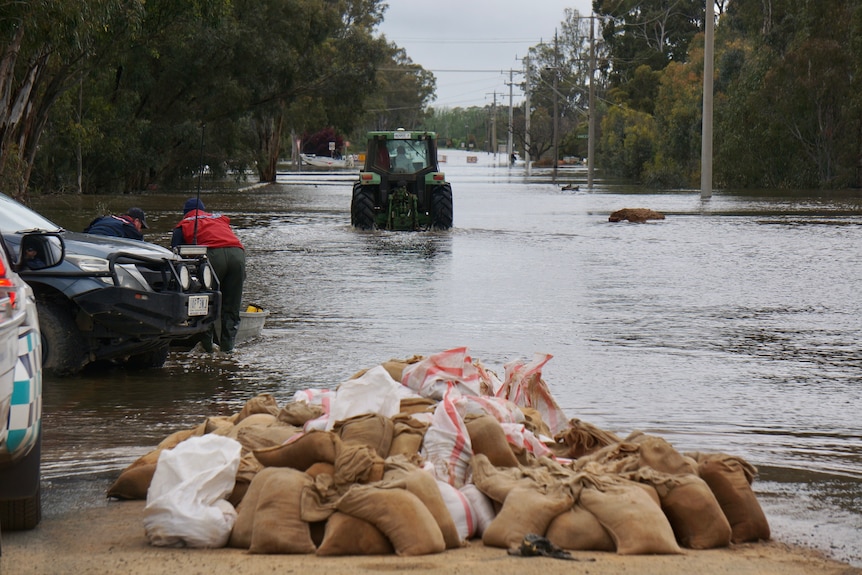 A tractor drives through floodwaters, with a pile of sandbags in the foreground.