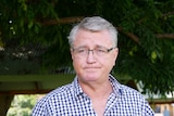 A man with grey hair glasses and a button up shirt looks off camera in an outdoor setting.