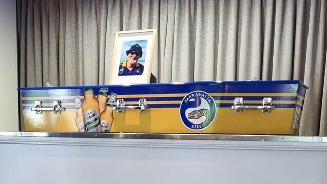 A coffin painted yellow and blue with the parramatta eels logo and beers on the side, photo on top. 