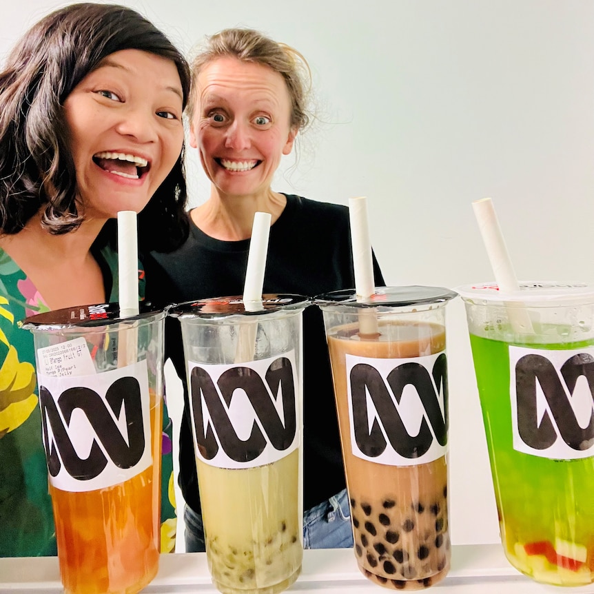 Karen Tong and Jessicah Mendes stand behind a row of four large cups of colourful bubble tea. 