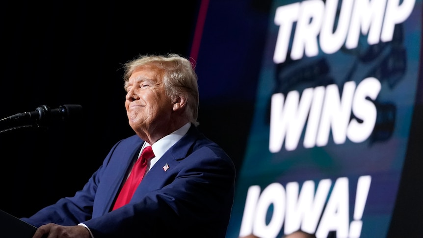 Donald Trump smiles on stage next to a sign saying TRUMP WINS IOWA