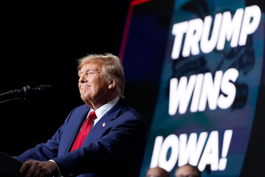 Donald Trump smiles on stage next to a sign saying TRUMP WINS IOWA