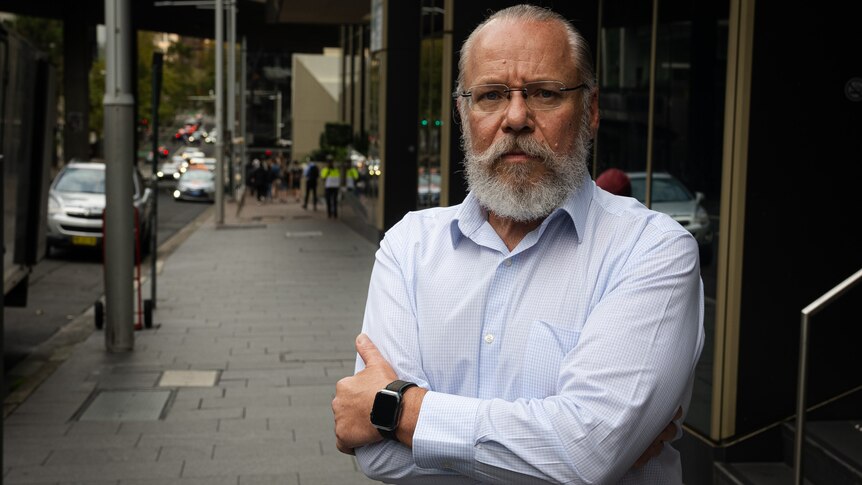 An older man wearing a collared shirt with a white beard stands on a footpath with his arms crossed.