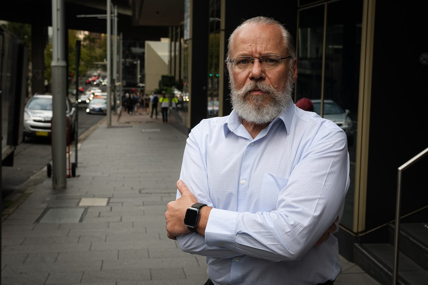 An older man wearing a collared shirt with a white beard stands on a footpath with his arms crossed.