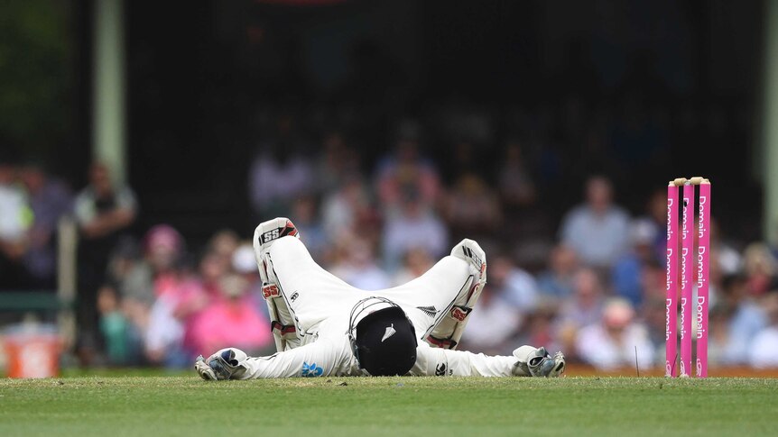 A wicketkeeper with his helmet on lies on the ground next to the stumps looking up at the sky.