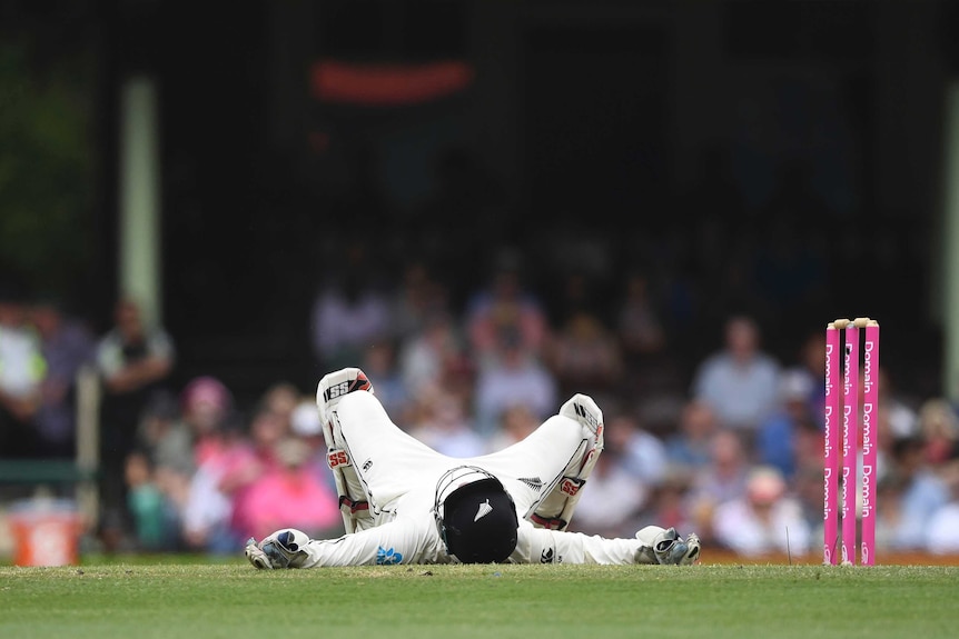 A wicketkeeper with his helmet on lies on the ground next to the stumps looking up at the sky.