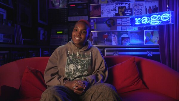an image of arlo parks sitting on the red rage couch smiling at the camera