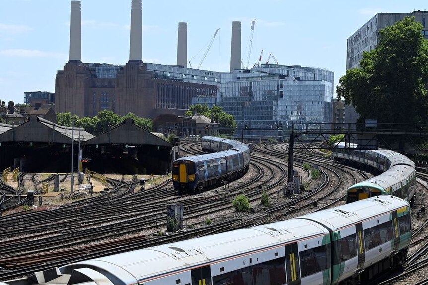 Two trains snake around a railway track in london on a bright and seemingly hot summer's day with factories and buildings behind