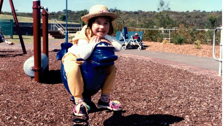 A girl smiles at the camera while sitting on playground equipment wearing a straw hat.