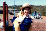 A girl smiles at the camera while sitting on playground equipment wearing a straw hat.