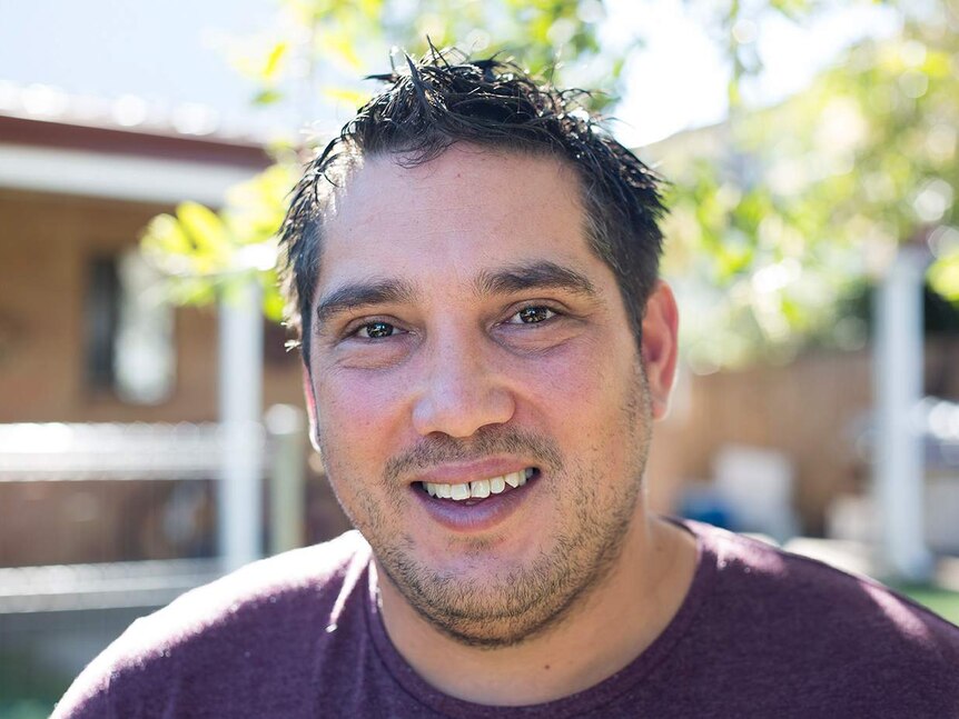 A man smiles at the camera. Wearing a purple t-shirt, with a five-o'clock shadow. Trees and house in background.