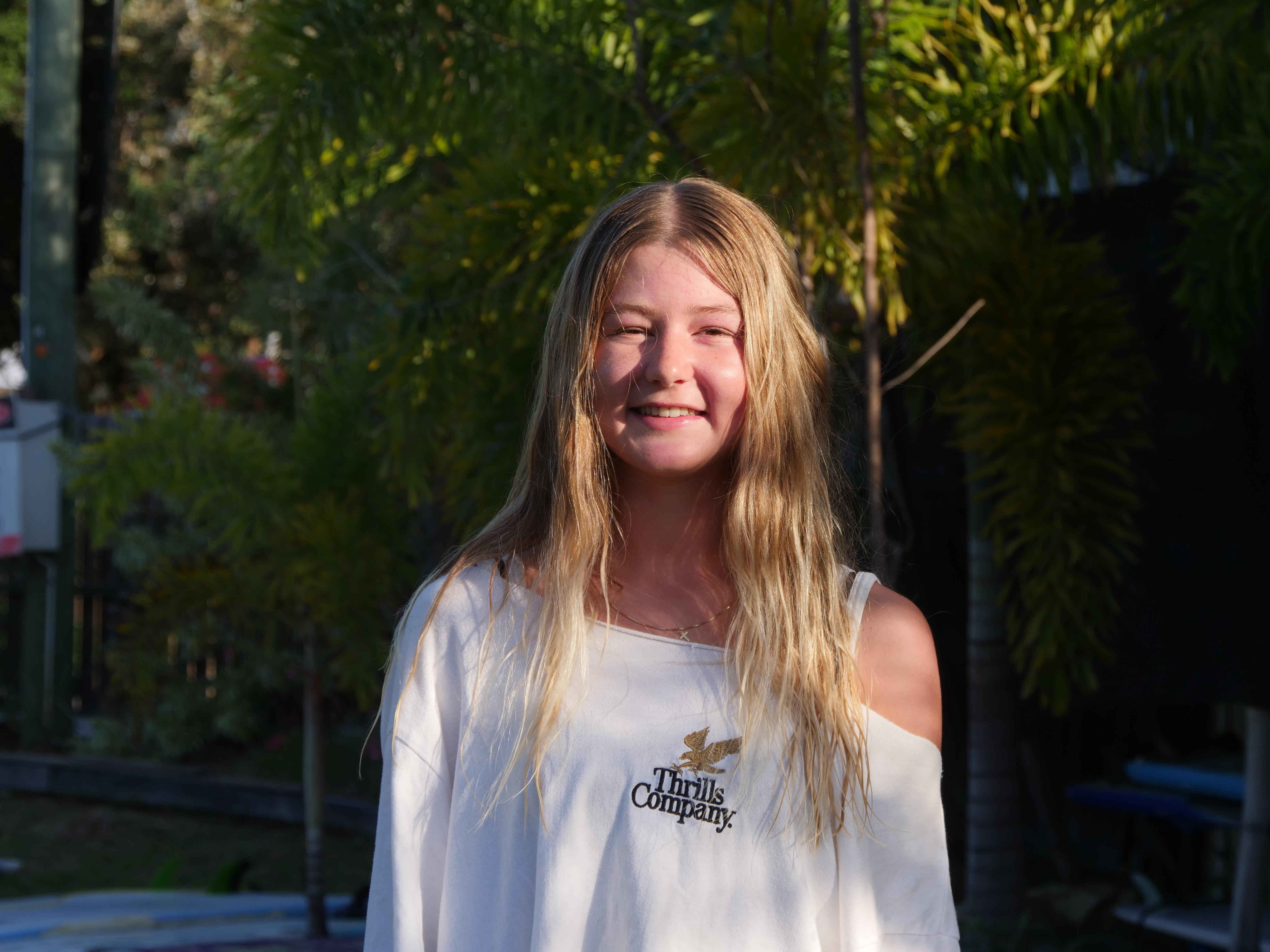 Una chica con cabello largo y rubio y una camiseta con hombros descubiertos sonríe directamente a la cámara y el sol brilla en su rostro.