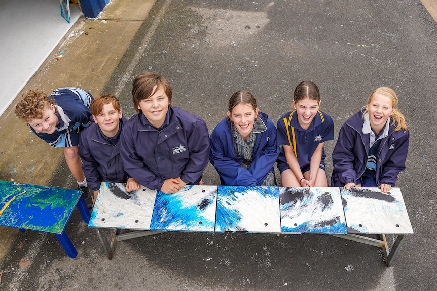 Six primary school aged children lean against a bright blue bench smiling.