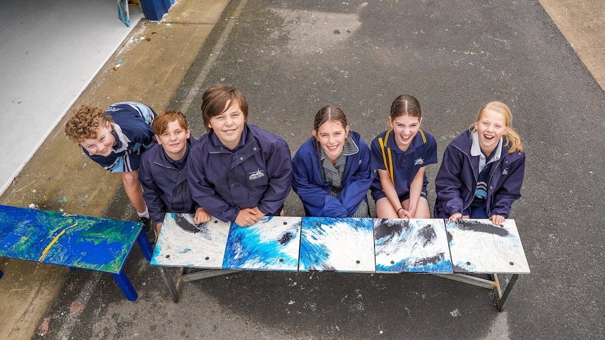 Six primary school aged children lean against a bright blue bench smiling.
