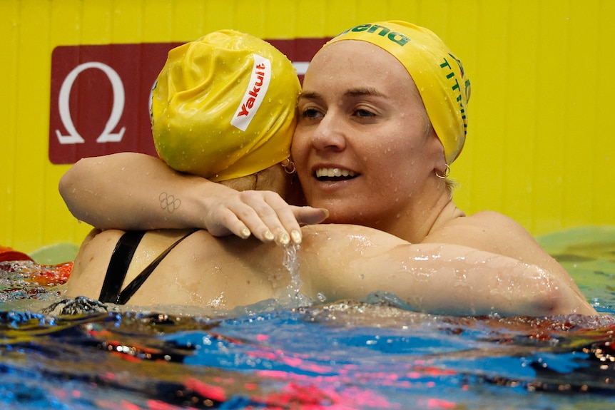 Two women hug in a pool wearing yellow swimming caps.