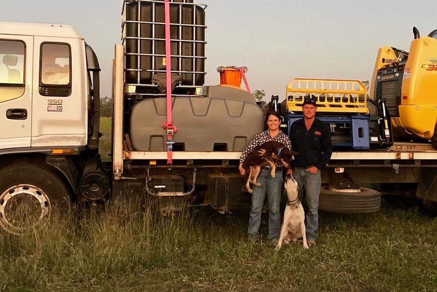 A man and woman stand in workwear in front of a truck of goods for building fences with two dogs.