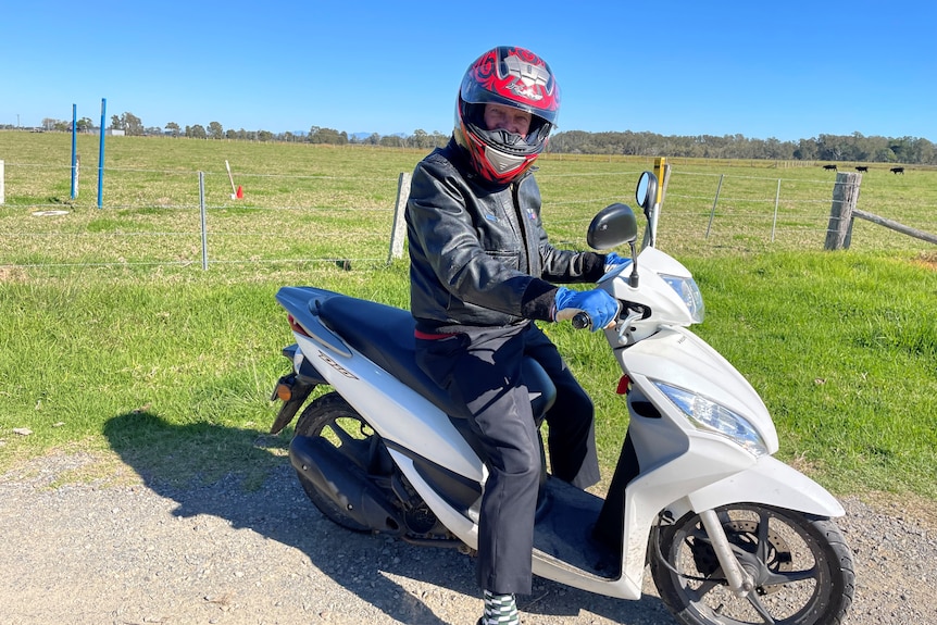 An older man on a motor scooter, wearing a jacket, long pants and a helmet, with a cow paddock behind him.