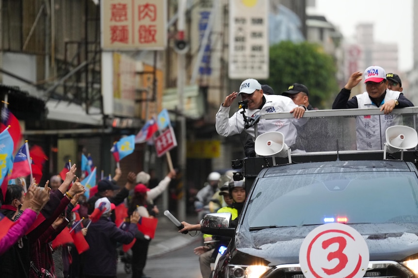 Two men stand on the back of a ute waving to people waving flags from the side of the road
