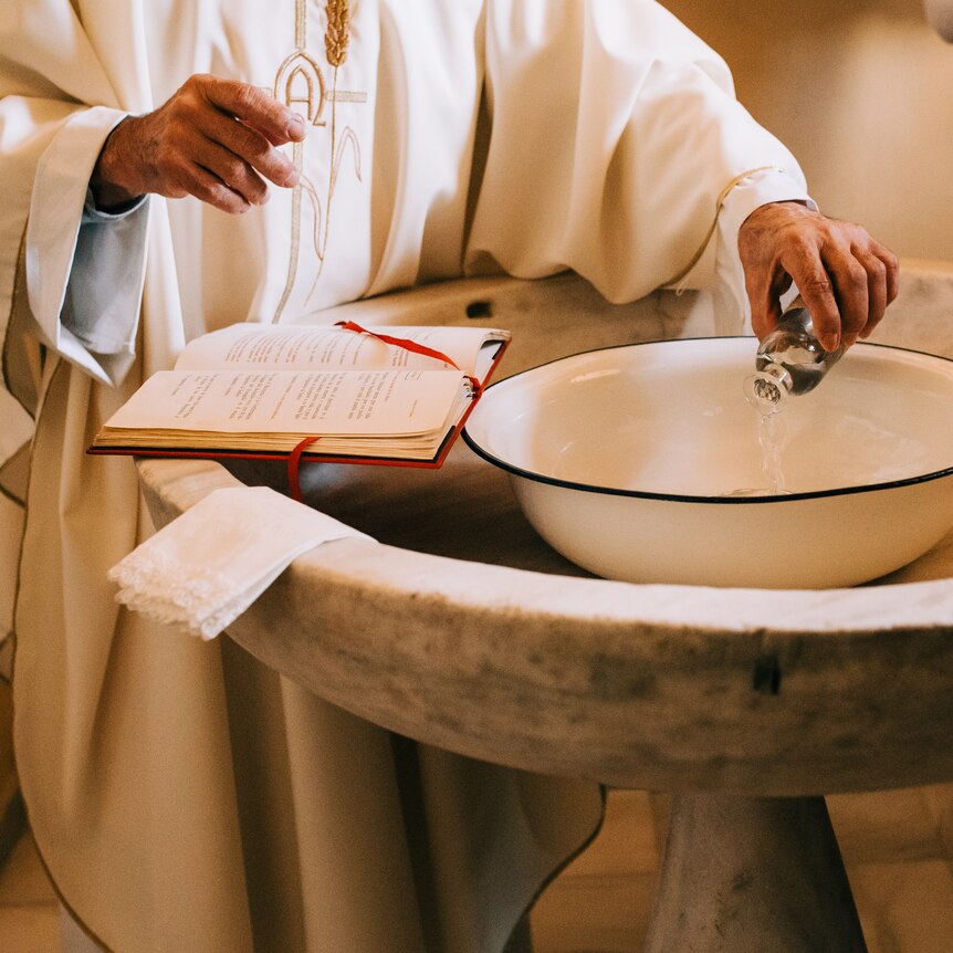 hands of priest in white robe pouring water into baptismal font