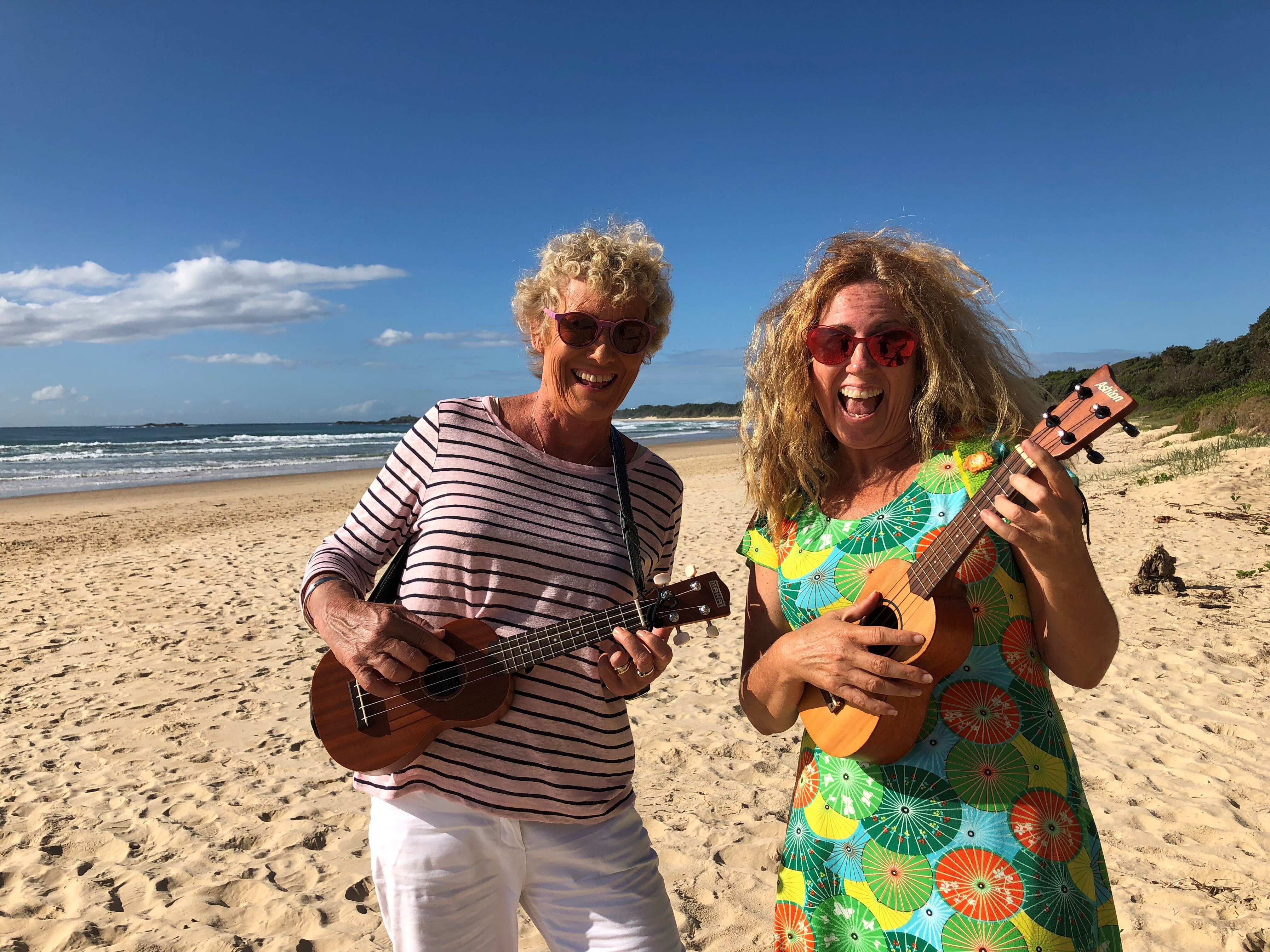 Two women laughing, wearing sunglasses, playing ukuleles on a white sand beach. 