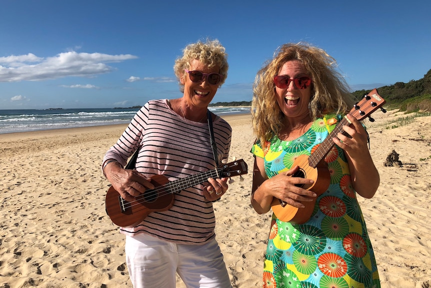Two women laughing, wearing sunglasses, playing ukuleles on a white sand beach. 