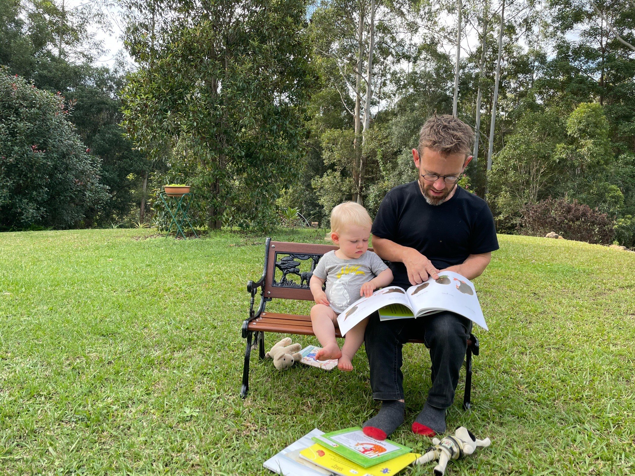 A man and a toddler sit together on a bench outside on grass as he reads to her from a book.