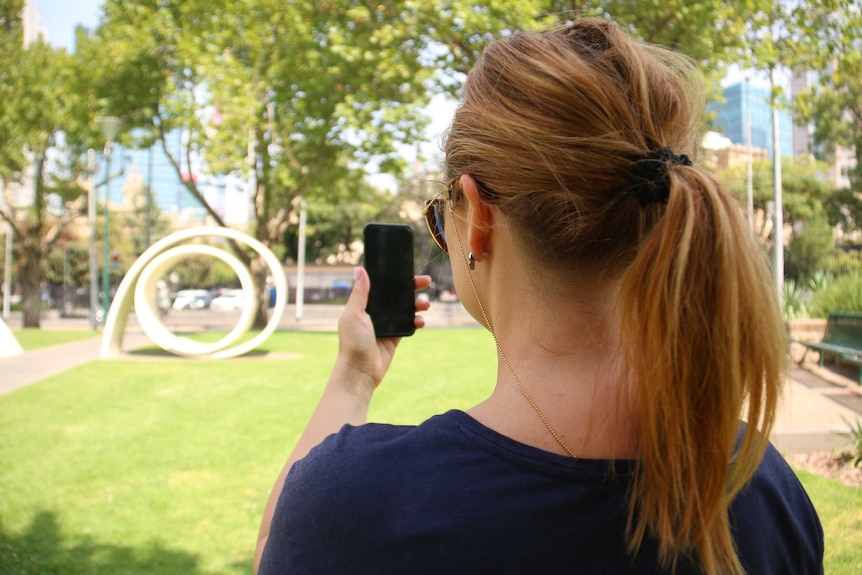 A woman sits in a park using her phone, her face is turned away from the camera.