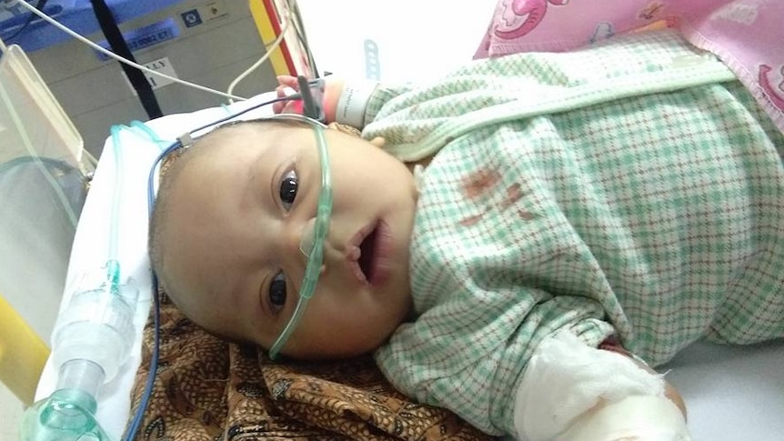 A baby lies in a hospital bed. His shirt is blood-stained and he his hooked up to an oxygen machine