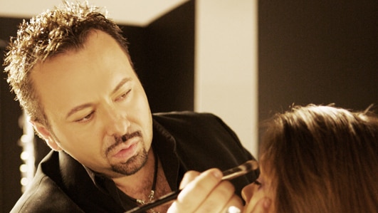 Napoleon Perdis stands with makeup brush in his hand as he applies eye shadow to a seated model.