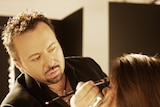 Napoleon Perdis stands with makeup brush in his hand as he applies eye shadow to a seated model.