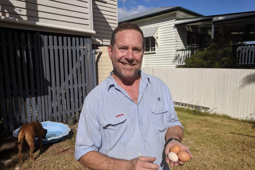Man standing in a suburban backyard, smiling, holding three eggs