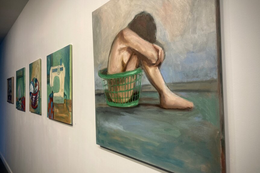Several paintings on a white wall, one in the forefront of a woman crouched in a washing basket. 