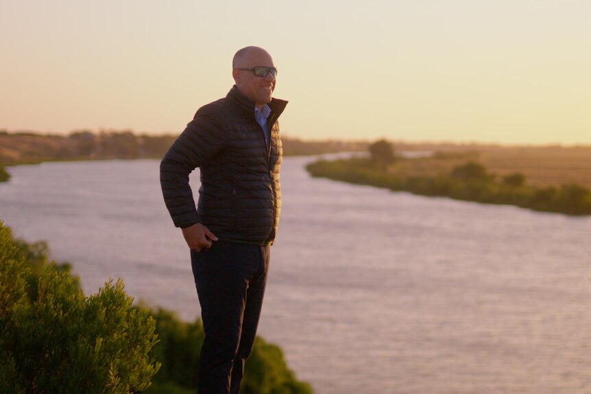 A bald man wears sunglasses, he wears a black puffer jacket black pants, he smiles looking over an expansive river at sunset