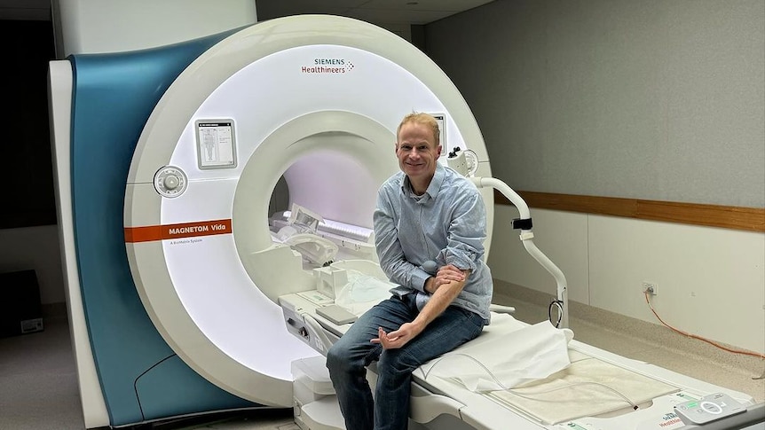 A man sits on the bed of an MRI machine looking at the camera.
