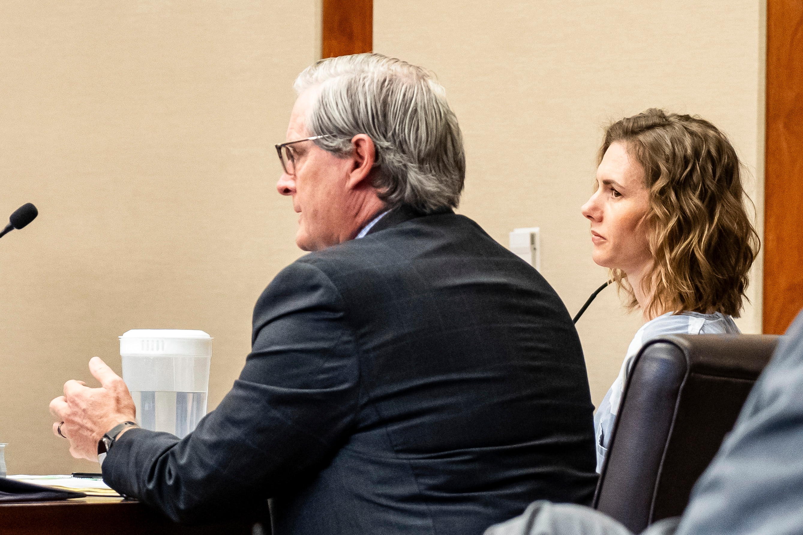 A woman with short, wavy, shoulder length brown hair in prison clothes blue and white striped sits before judge