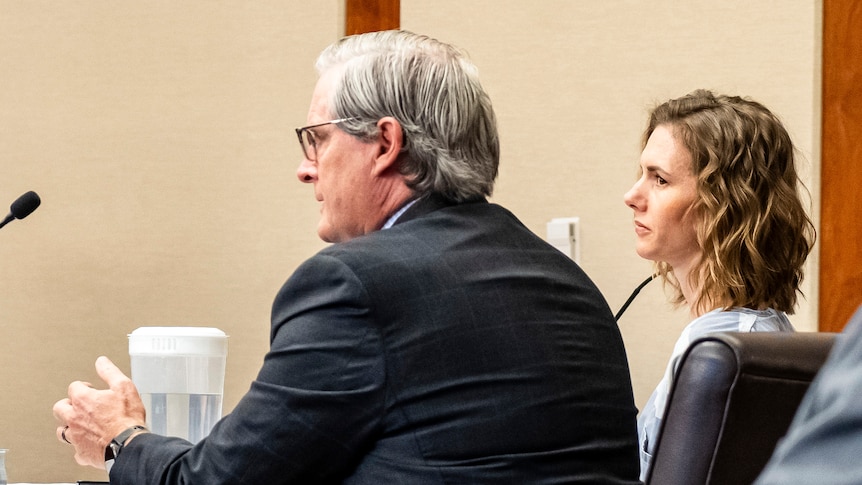 A woman with short, wavy, shoulder length brown hair in prison clothes blue and white striped sits before judge