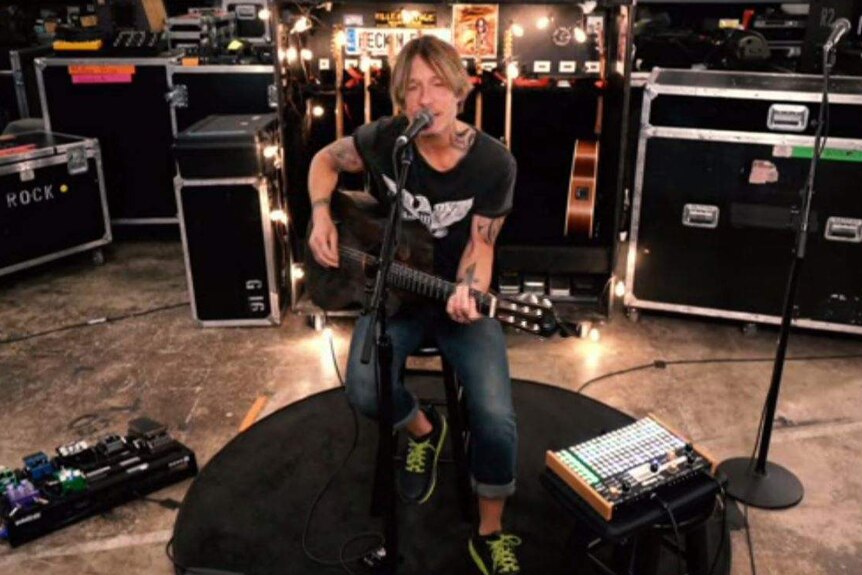 Keith Urban playing a guitar in a black shirt with guitars and musical equipment behind him