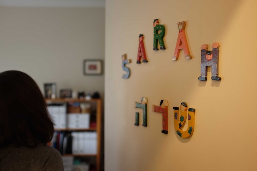 A woman enters her room, the door decorated with her name, Sarah, in wooden figurines.