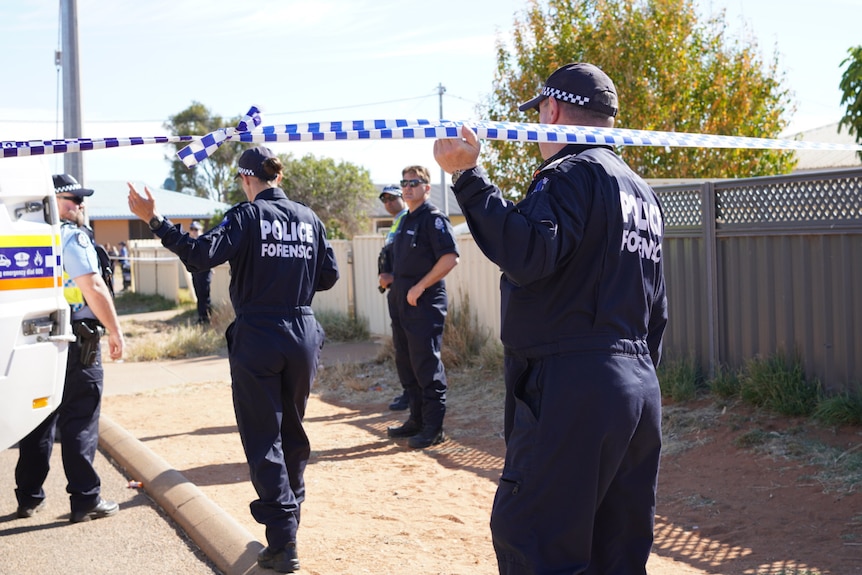 A police forensic officer ducks under police tape and walks towards other officers outside a house.