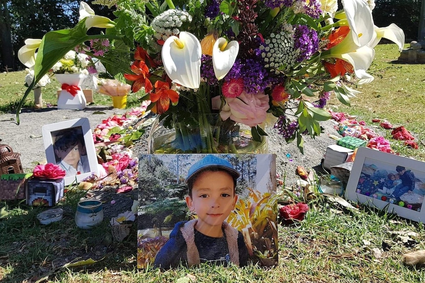 A grave adorned with brilliant flowers and various ornaments with a picture of a young boy in a hat sitting in front of it.