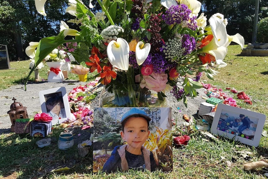 A grave adorned with brilliant flowers and various ornaments with a picture of a young boy in a hat sitting in front of it.