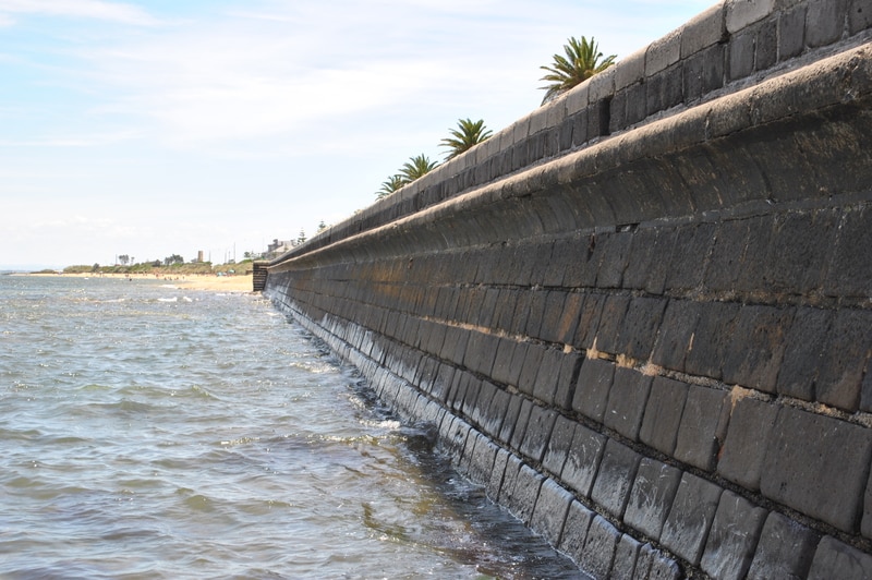 A bluestone seawall showing seawater and the beach in the background.