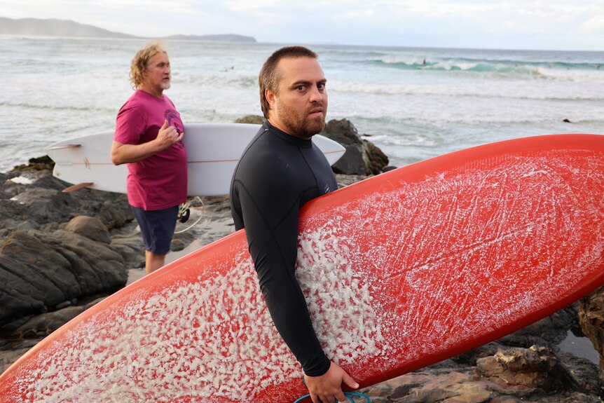 A man in a black top holds a red surfboard under his arm and another man in a purple top holds a white board with ocean behind t