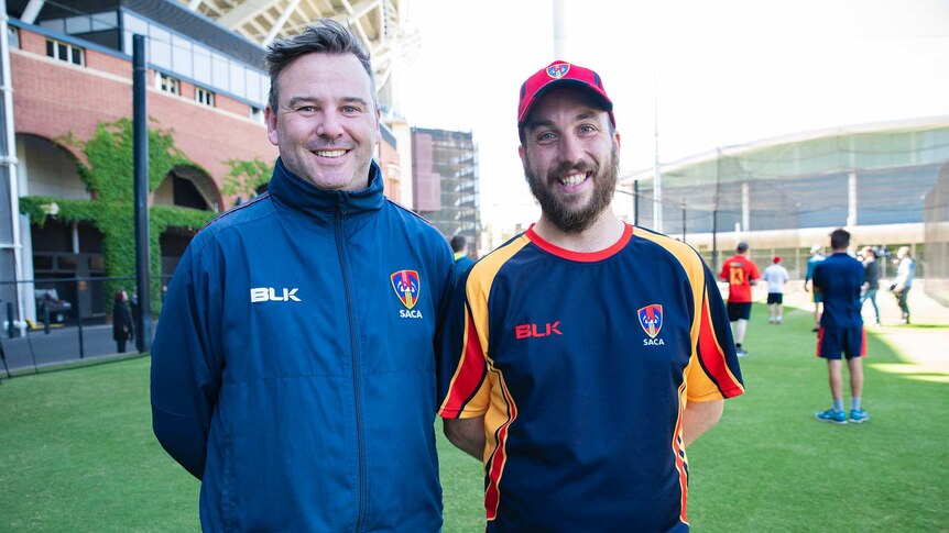 Two men — one older, the other apparently in his 20s — smile for a photo outside a sports venue.