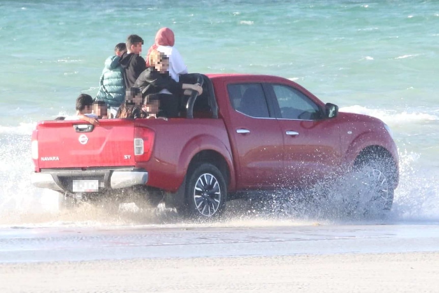 A red ute driving through the water near the beach with people in the back tray