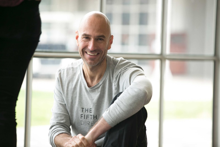 image of caucasian man grinning and looking forwards, wearing casual clothes and posing with one arm draped over his knee